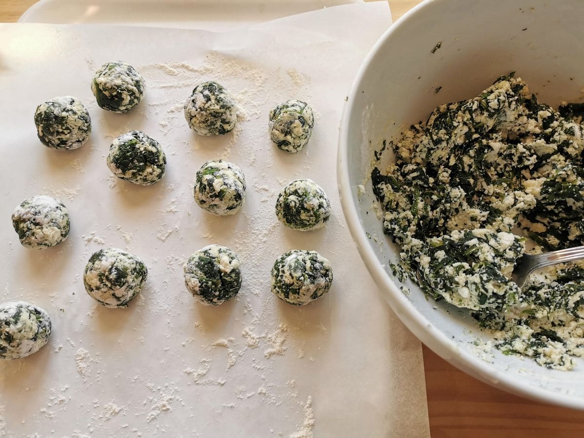Uncooked gnudi mixture in white bowl next to some ready rolled gnudi on flour dusted white surface.