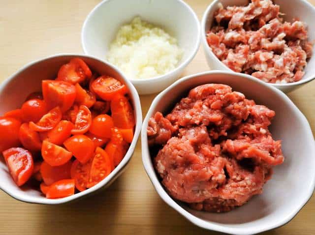 cut tomatoes, chopped onions, chopped sausage meat and ground beef in separate white bowls.