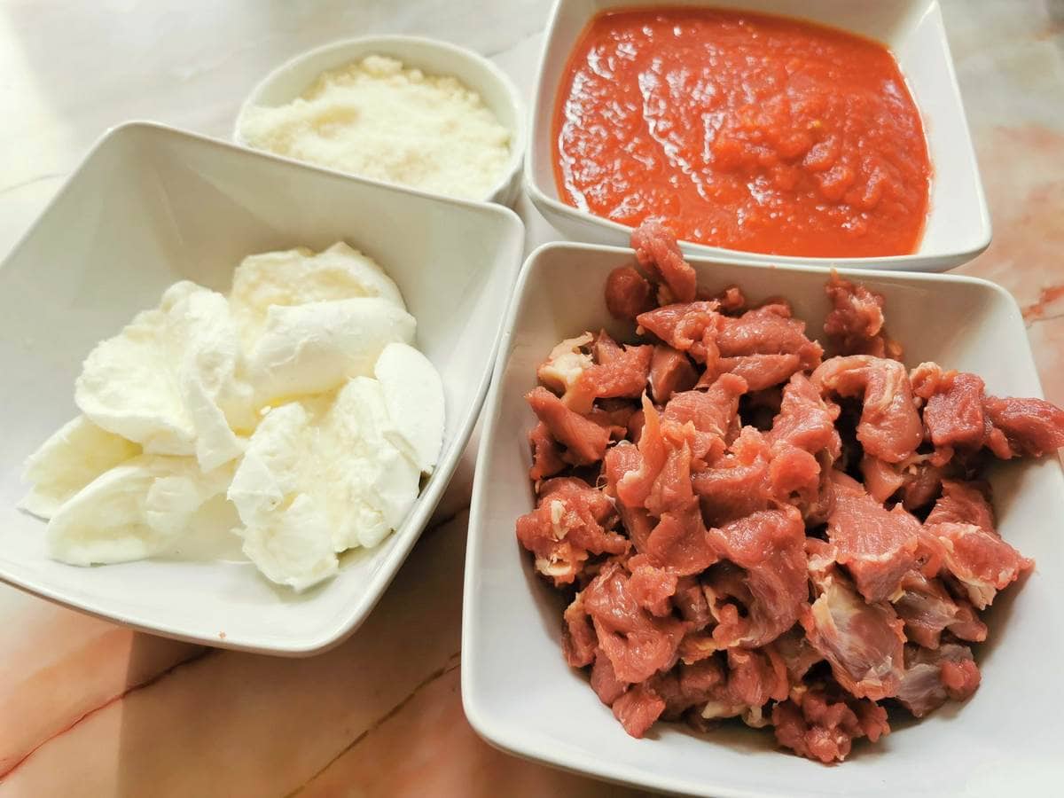 Sliced mozzarella in white bowl. Diced lamb in white bowl. Grated pecorino in white bowl and tomato passata in white bowl.