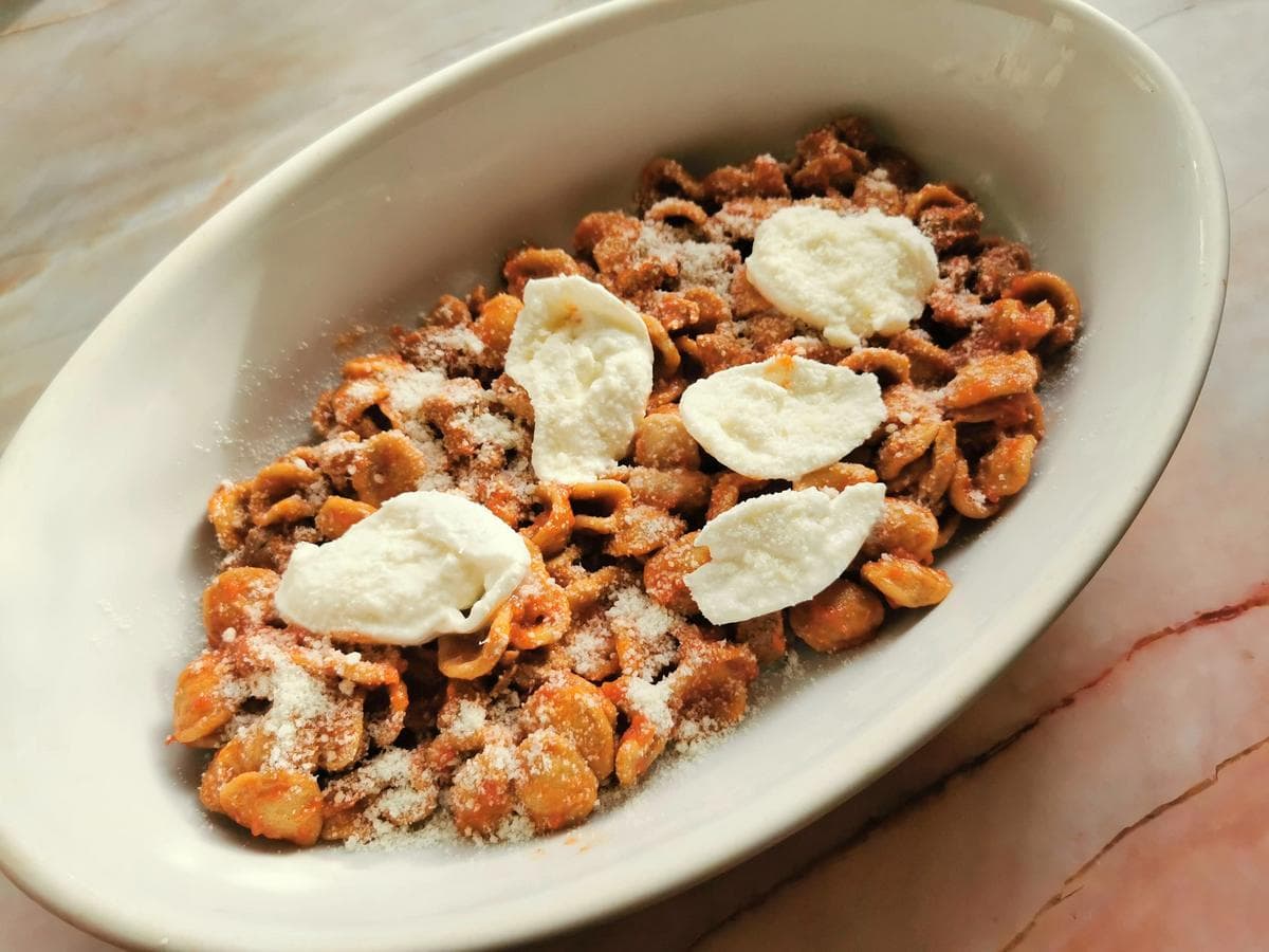 Slices of mozzarella and grated pecorino on top of orecchiette and lamb sauce in oval oven dish.