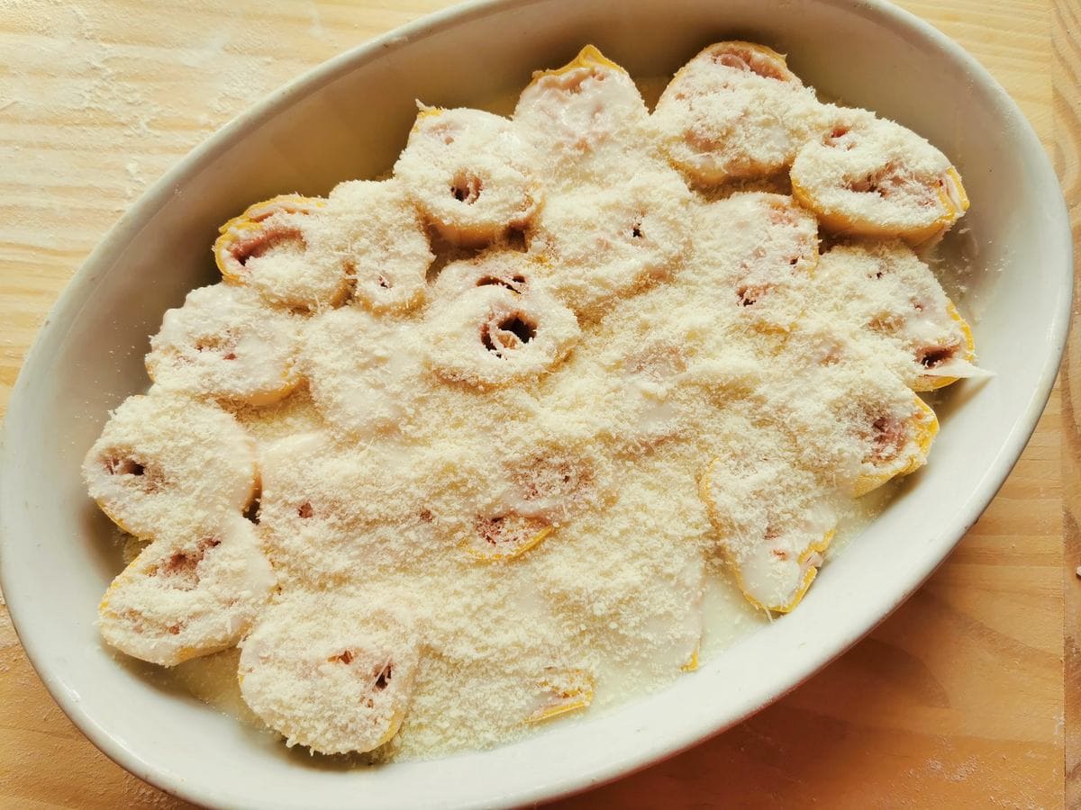 Uncooked pasta roses in white oval oven dish with béchamel and grated Parmigiano.