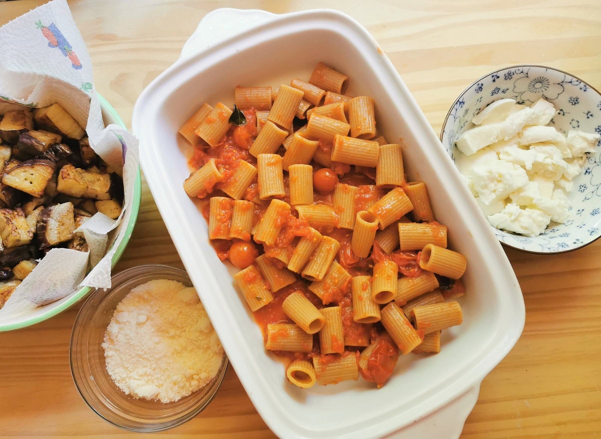 White oven dish with layer of whole wheat pasta and tomato sauce inside and fried eggplant cubes in a small bowl and cubes of mozzarella in a small bowl.