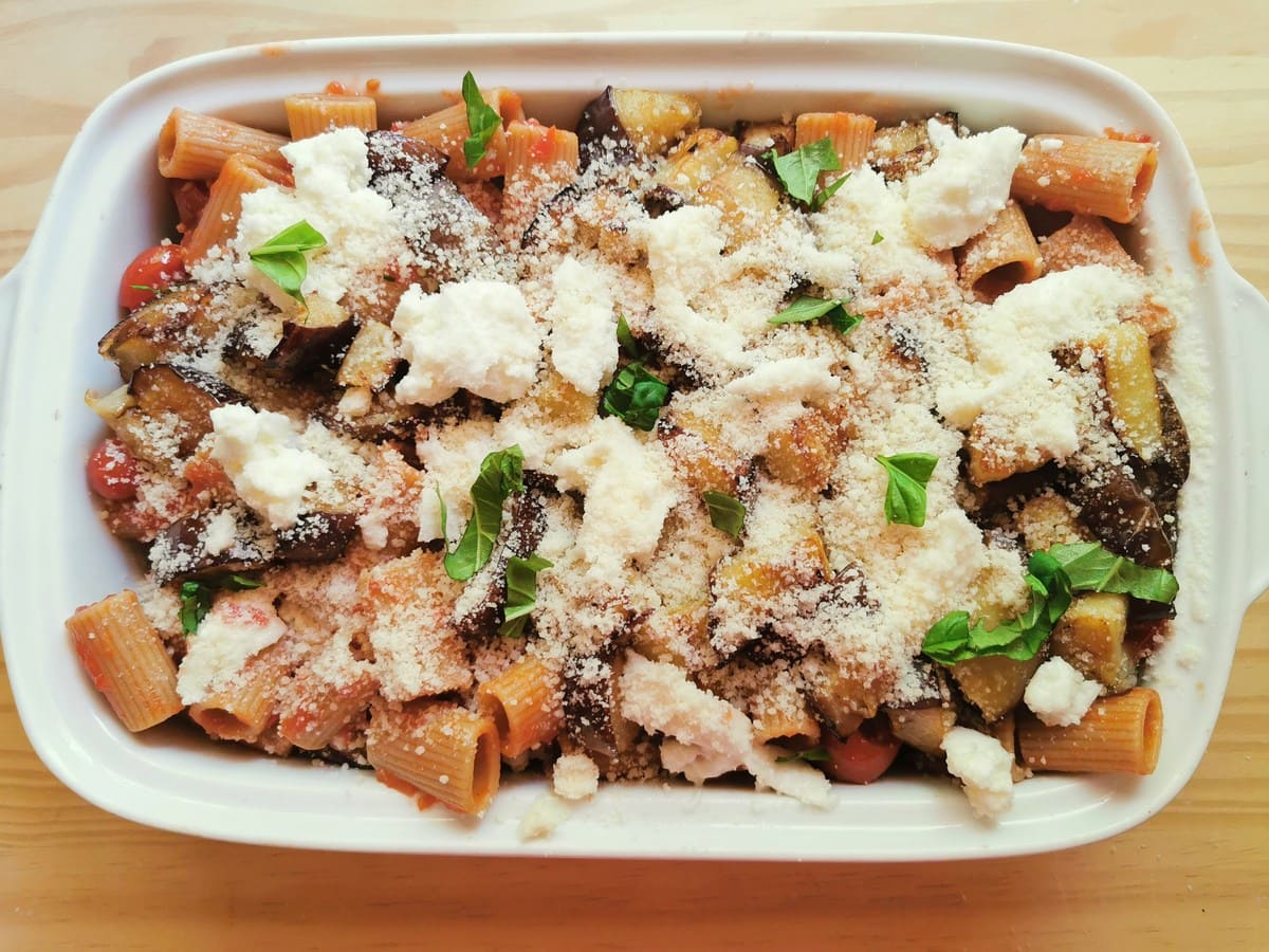 Whole wheat pasta with tomato sauce, fried eggplant, mozzarella and grated Parmigiano, topped with fresh basil leaves in white oven dish.