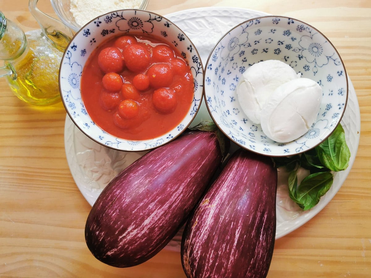 Ingredients for pasta with eggplant alla Siciliana sauce; 2 eggplants, mozzarella balls in small bowl, canned peeled cherry tomatoes in small bowl, basil leaves and olive oil in glass jug.