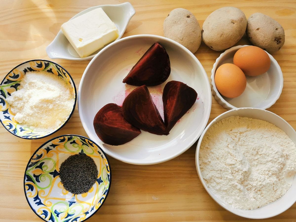 Ingredients for beetroot ravioli on wood worktop; flour, eggs, potatoes, butter, grated grana, poppy seeds and cooked beetroot.