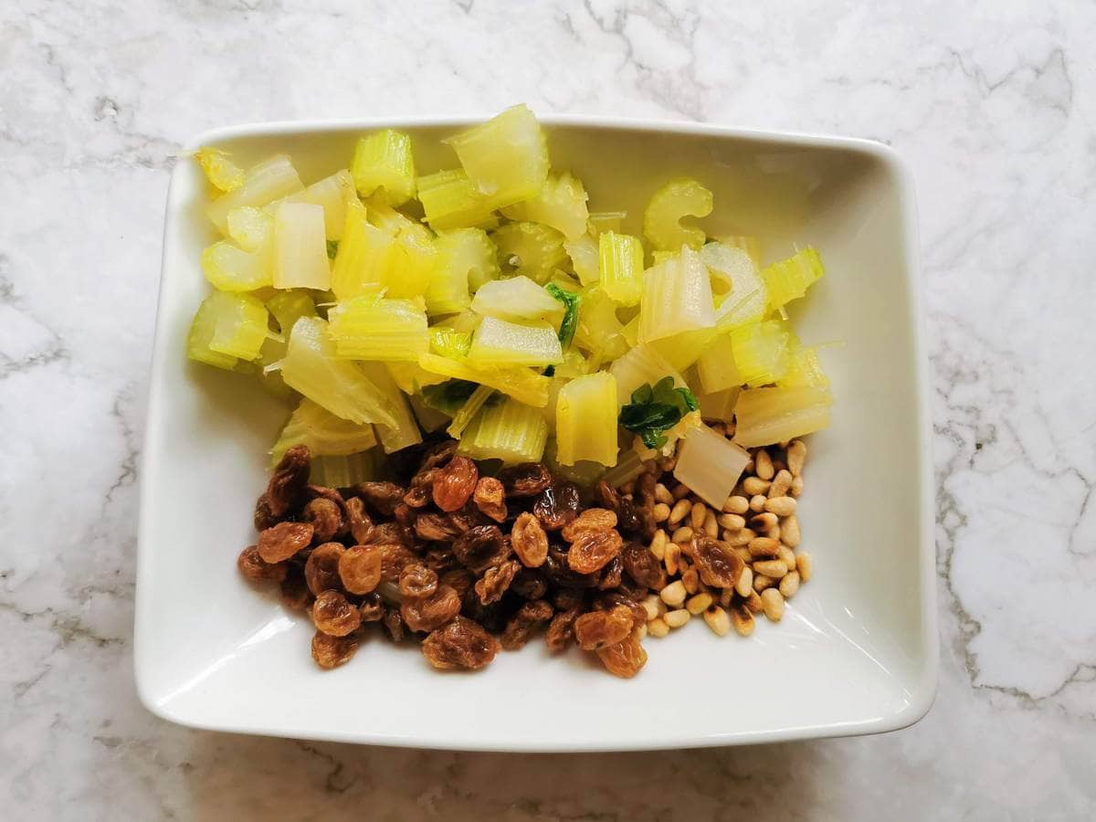 Soaked raisins, toasted pine nuts and boiled celery pieces in white bowl.