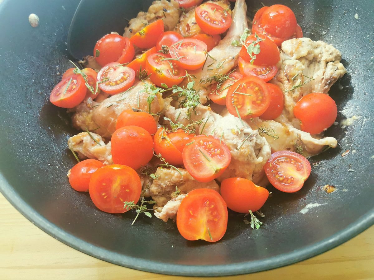 Halved cherry tomatoes and fresh herbs added to rabbit pieces cooking in deep skillet.