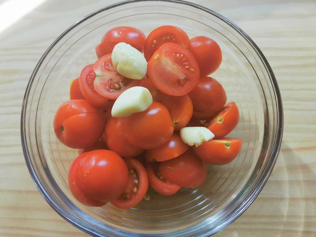Halved cherry tomatoes and peeled garlic cloves in glass bowl.