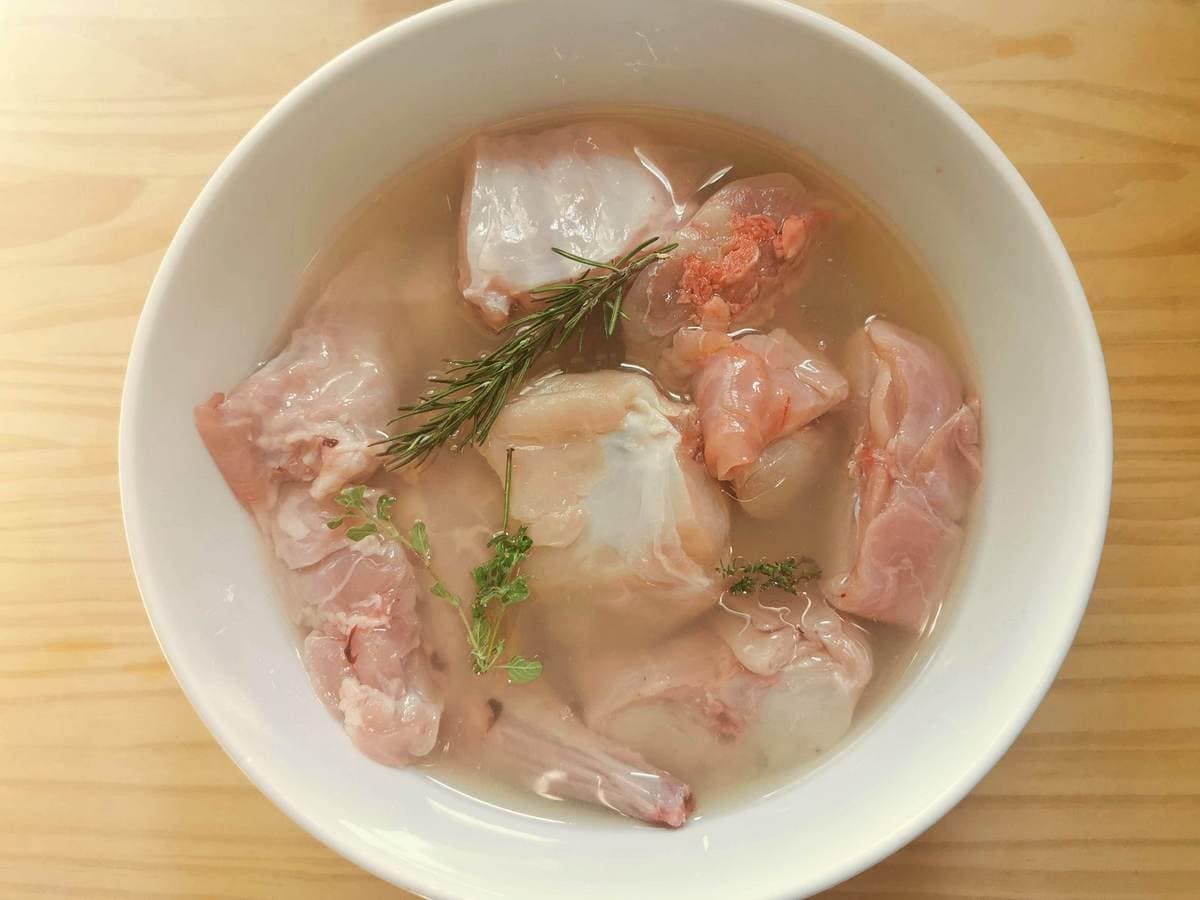 Pieces of rabbit marinating in white wine with herbs in a white bowl.