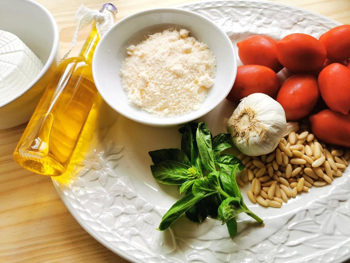 Ingredients for Sicilian pesto on white plate. These include ripe tomatoes, parmesan, fresh ricotta, pine nuts, garlic, basil and extra virgin olive oil.