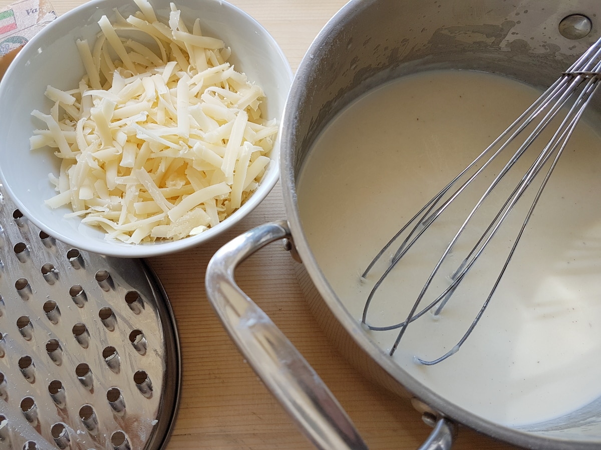 Bechamel sauce in saucepan and grated cheese in white bowl.