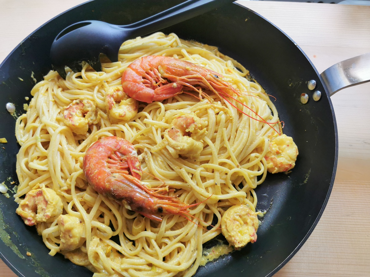 Linguine mixed with peeled and unpeeled cooked prawns in saffron sauce in skillet.