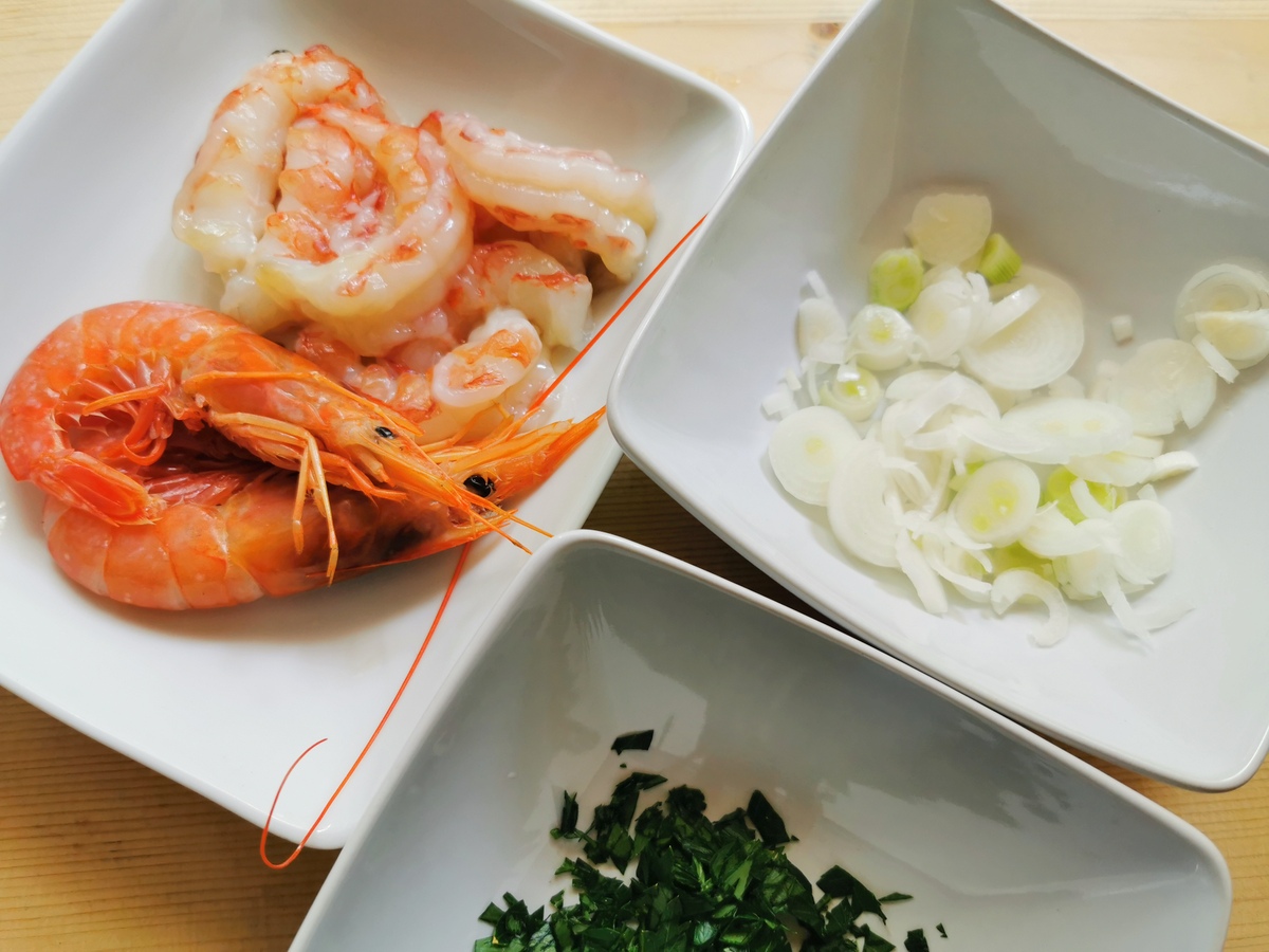 Sliced spring onion, chopped parsley and peeled prawns in separate white bowls.