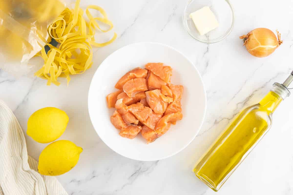 Salmon, lemon, tagliatelle, onion, butter and extra virgin olive oil on a kitchen counter.