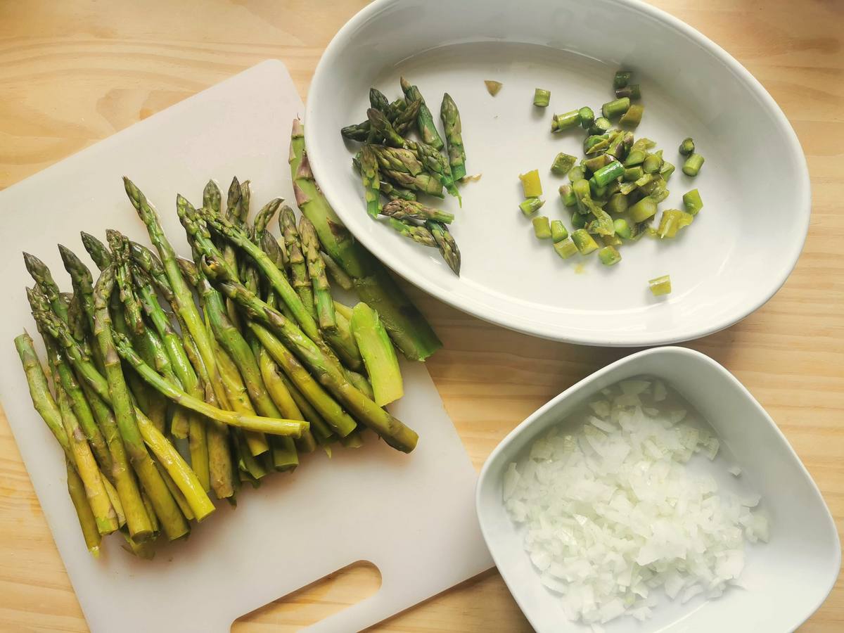 Blanched asparagus on white chopping board, peeled and chopped onion in white bowl and cut asparagus spears and stalks in white bowl.