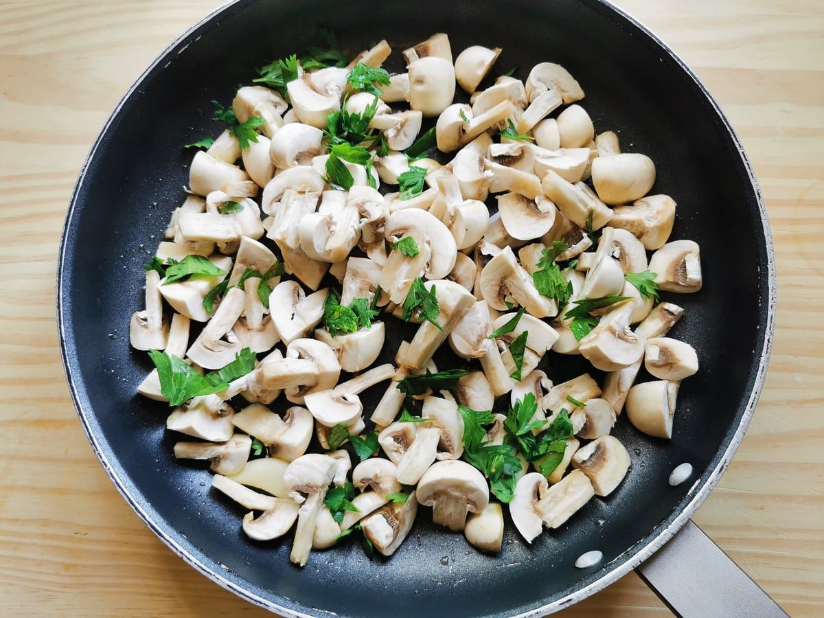 Sliced button mushrooms in skillet with garlic cloves and chopped parsley.