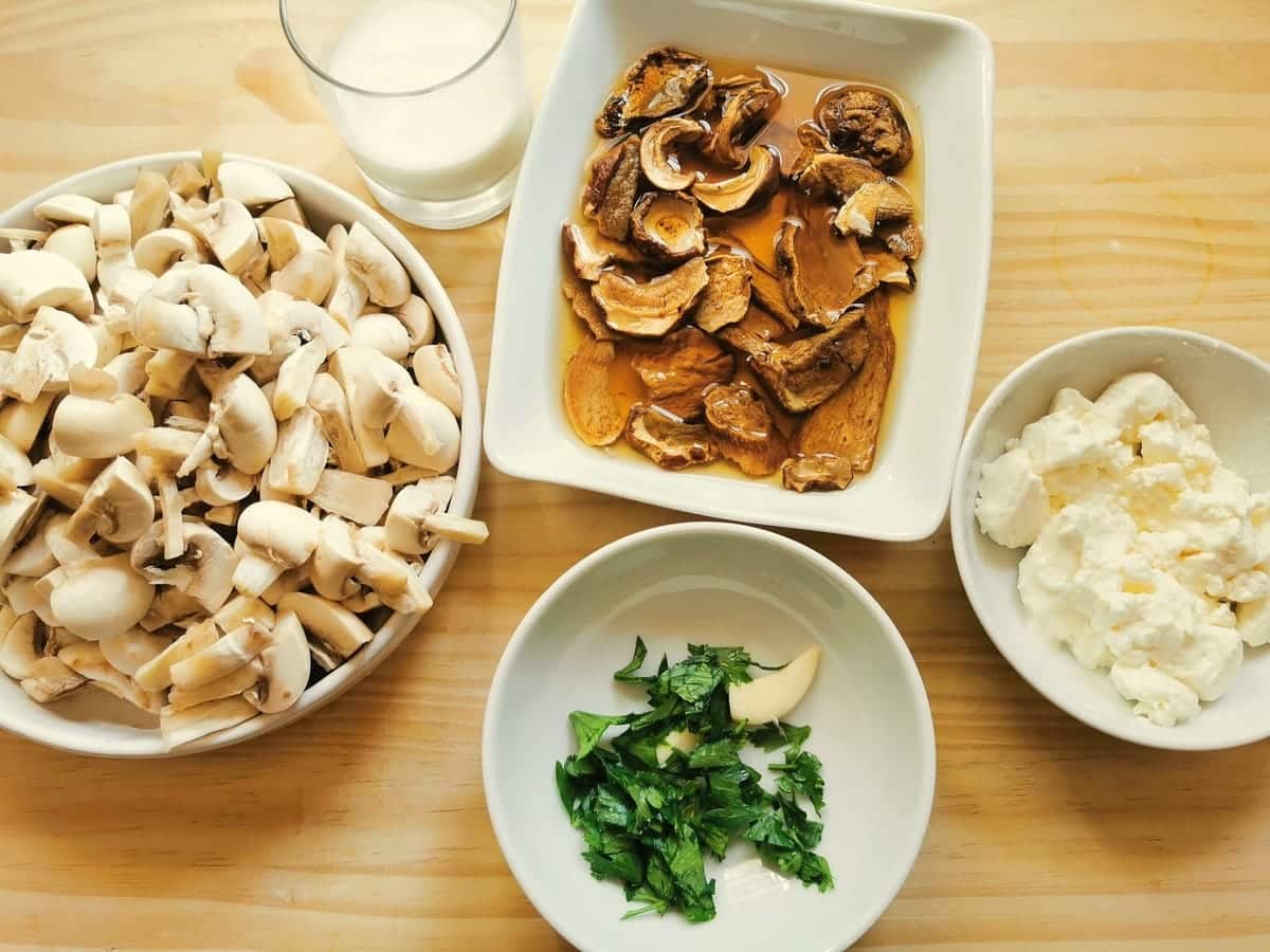 Sliced button mushrooms in white bowl. Dried porcini soaking in white bowl. Chopped parsley and peeled garlic in white bowl and stacchino in white bowl.