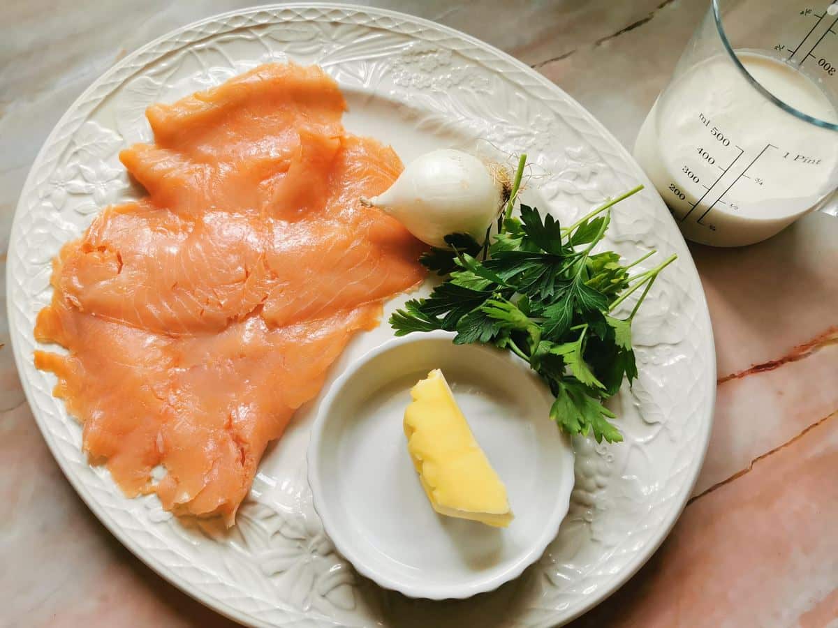 Ingredients for smoked salmon pasta. Smoked salmon, cream, onion, butter and parsley all on white plate.