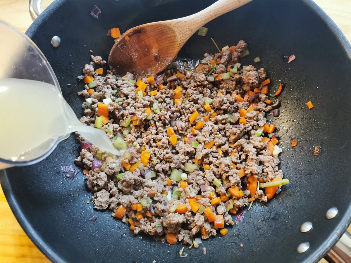 vegetable stock being poured into beef ragu in skillet.