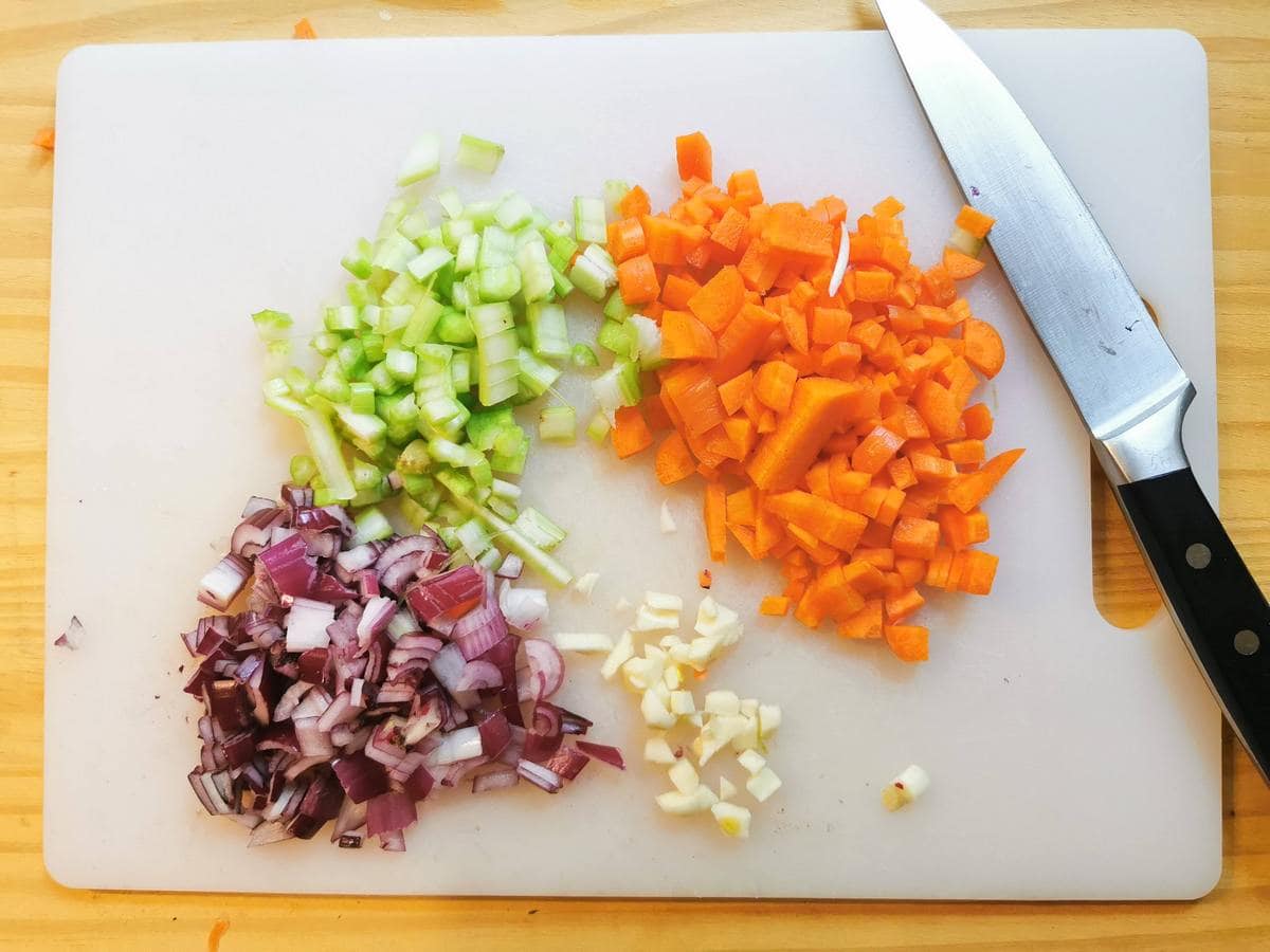 Chopped onion, garlic, carrots and celery on white chopping board with knife.