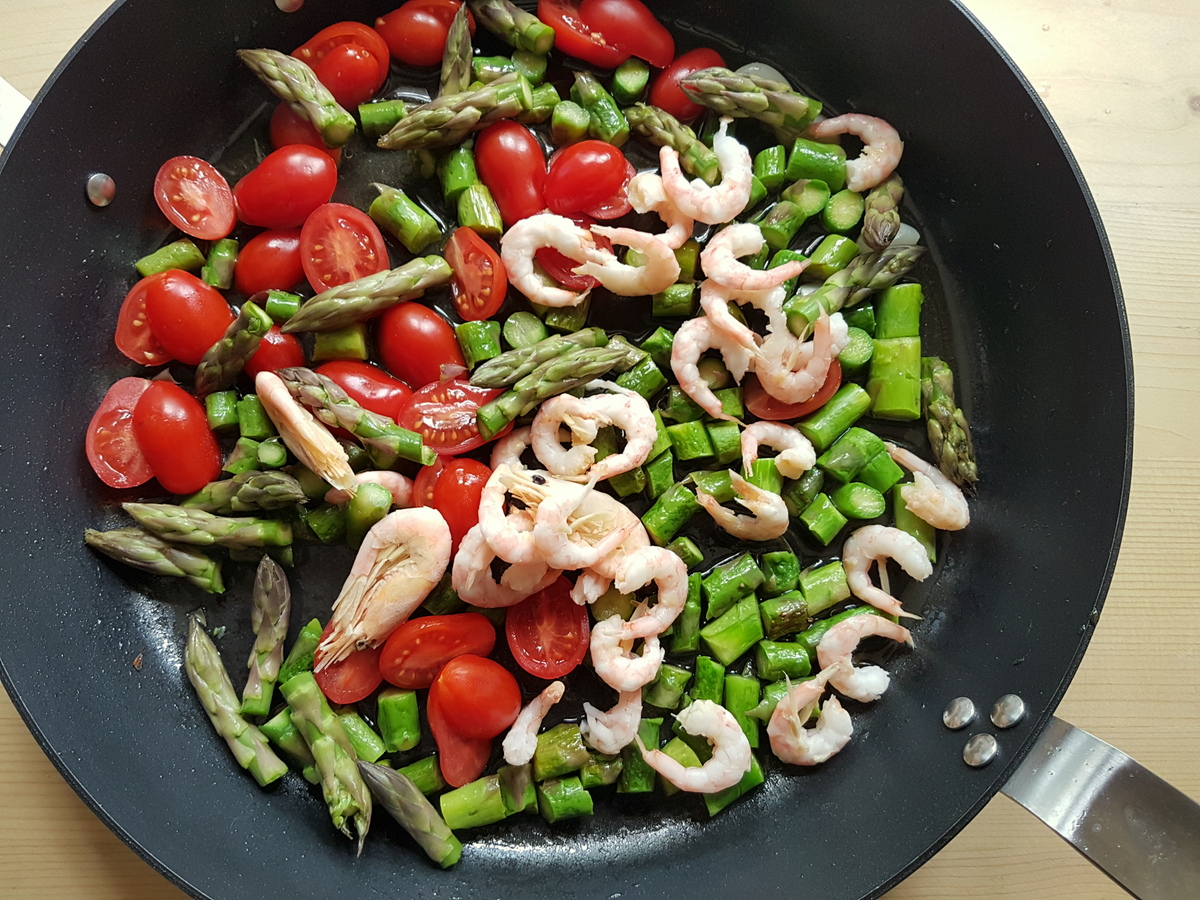 Cut asparagus stalks, asparagus spears, datterini tomatoes, garlic cloves and peeled shrimp cooking in skillet.