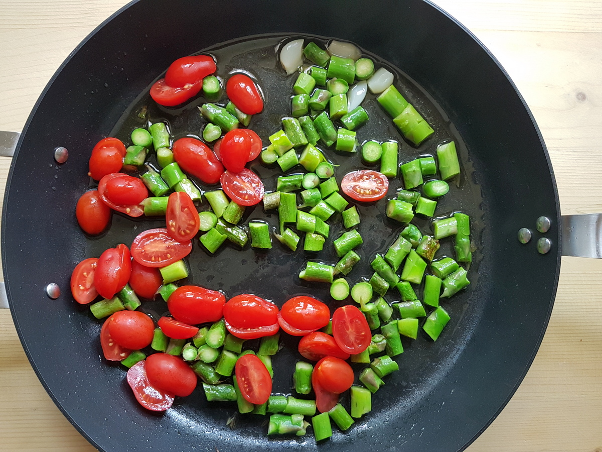 Cut asparagus stalks, whole garlic cloves and halved datterini tomatoes cooking in skillet.