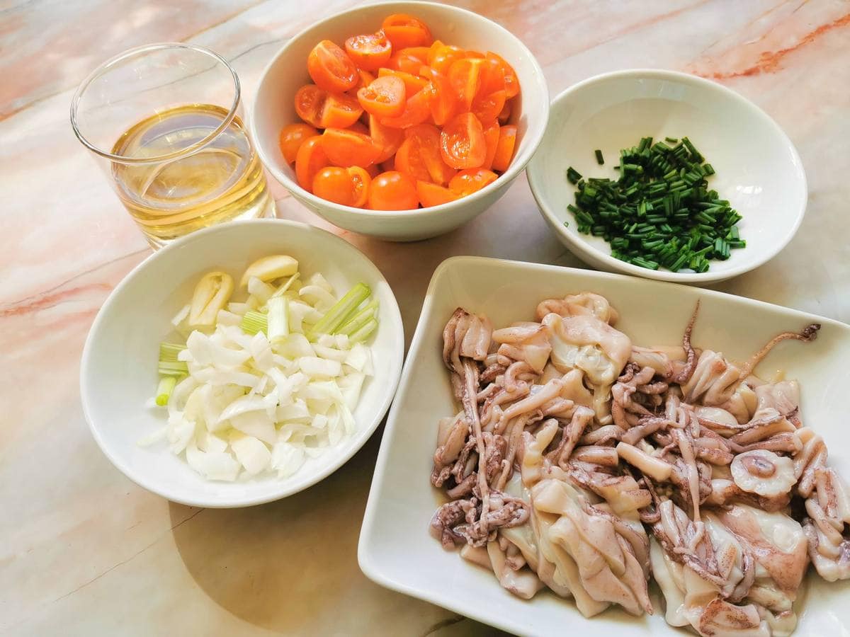 chopped spring onion in white bowl. Quartered cherry tomatoes in white bowl. Chopped chives in white bowl. Cleaned and cut squid in white bowl.