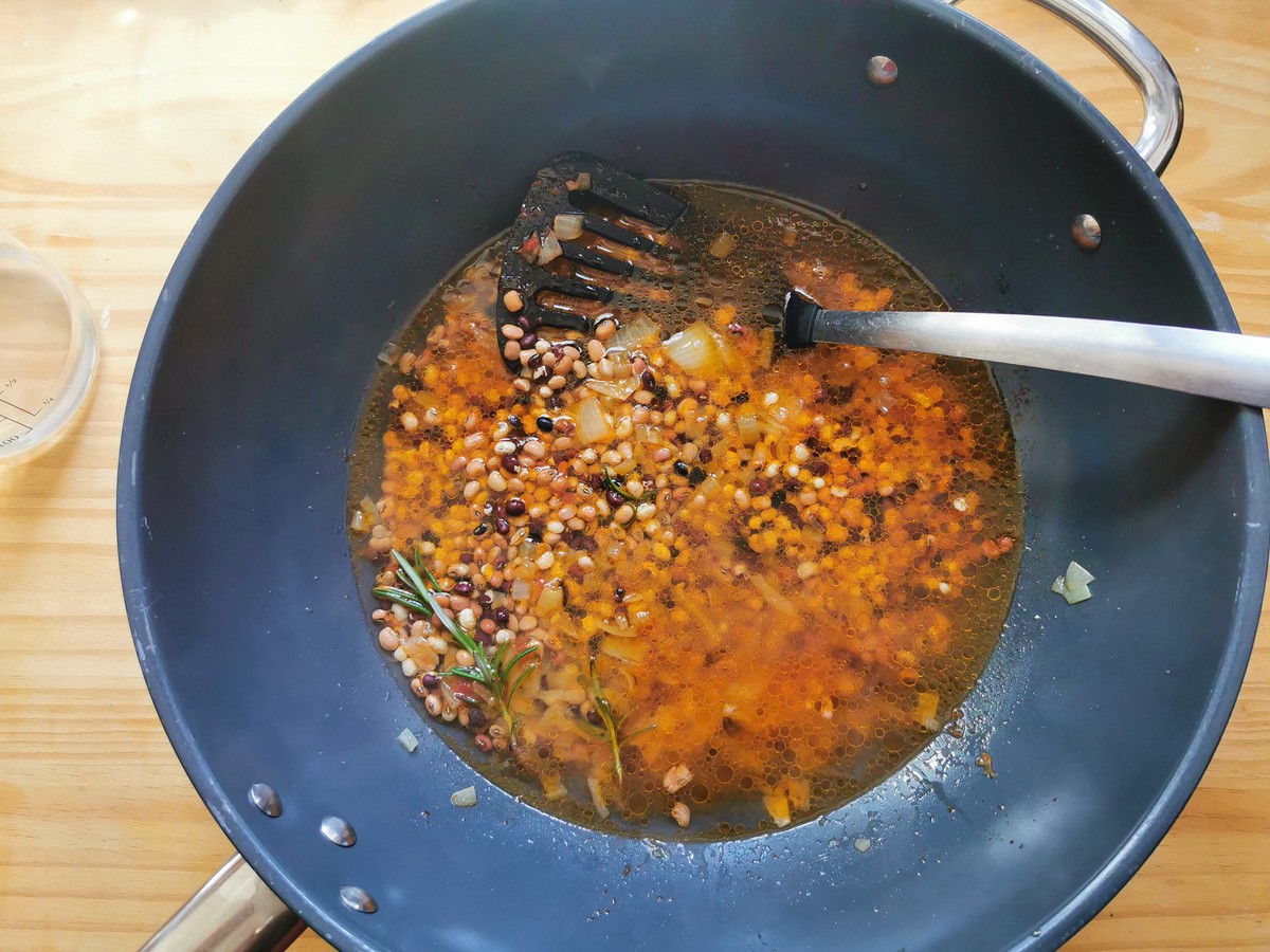 Uncooked Trasimeno beans with onion, tomato paste and rosemary sprigs covered with water in deep frying pan.