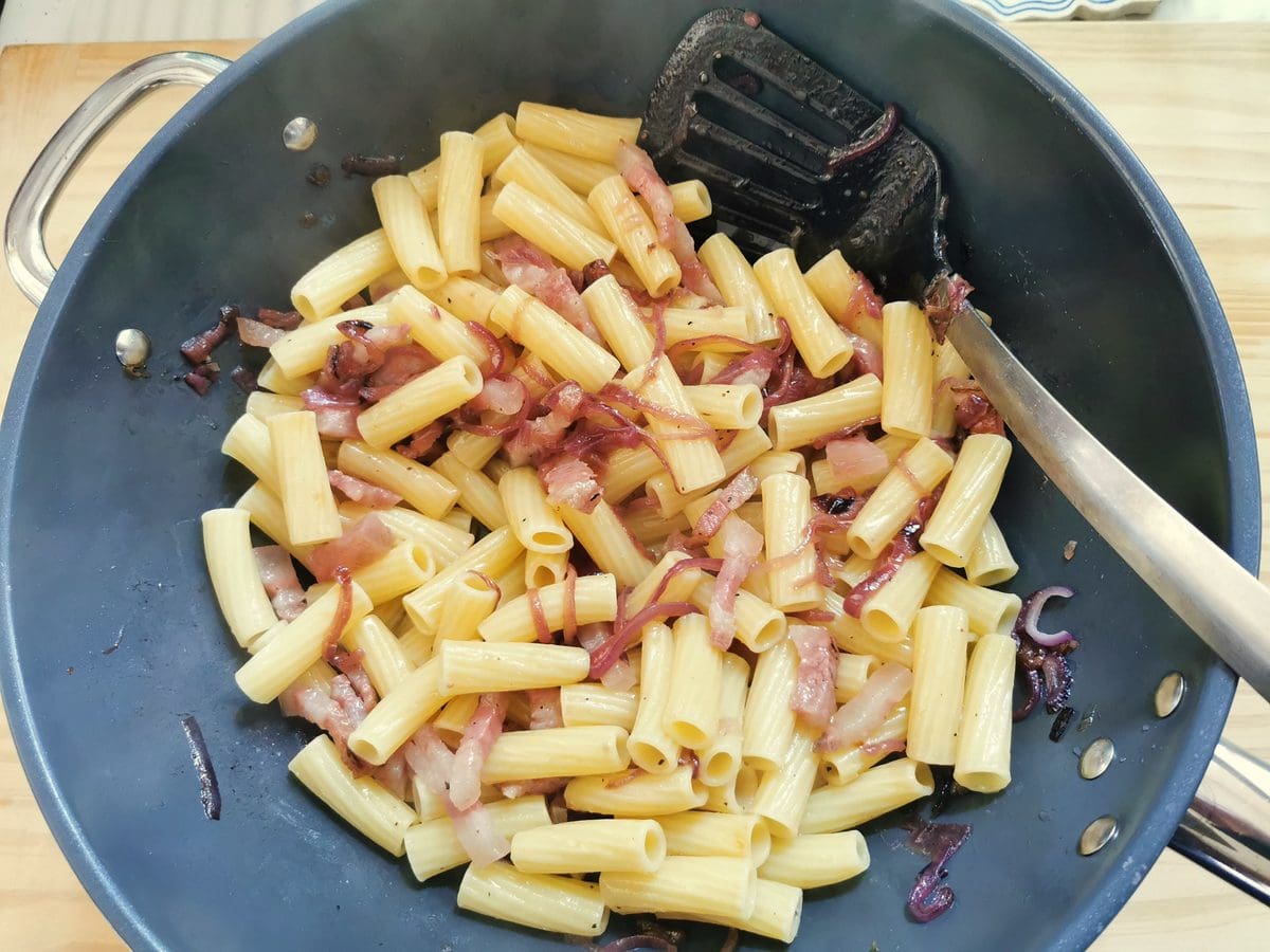 Cooked rigatoni mixed with red onion and guanciale in large skillet.
