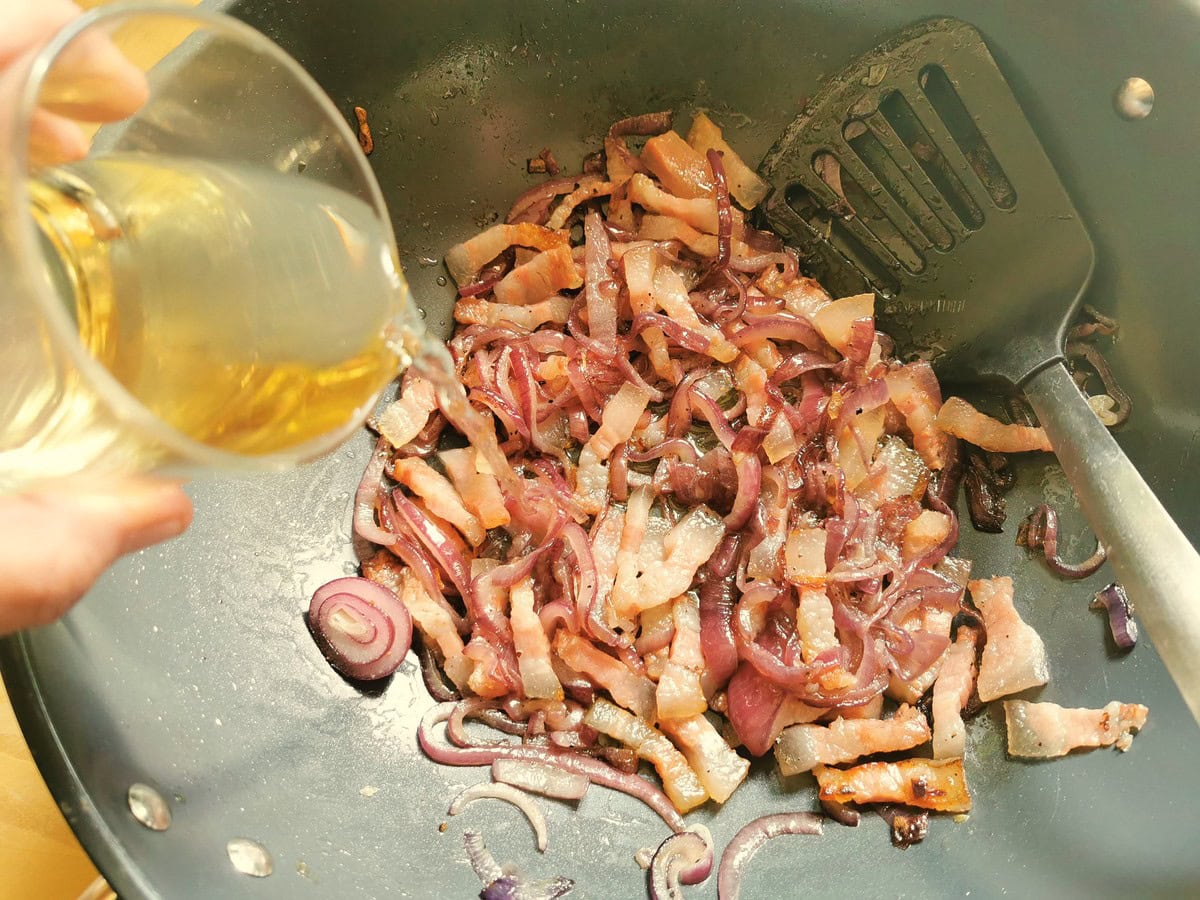 White wine being poured into skillet with red onion and guanciale lardons.