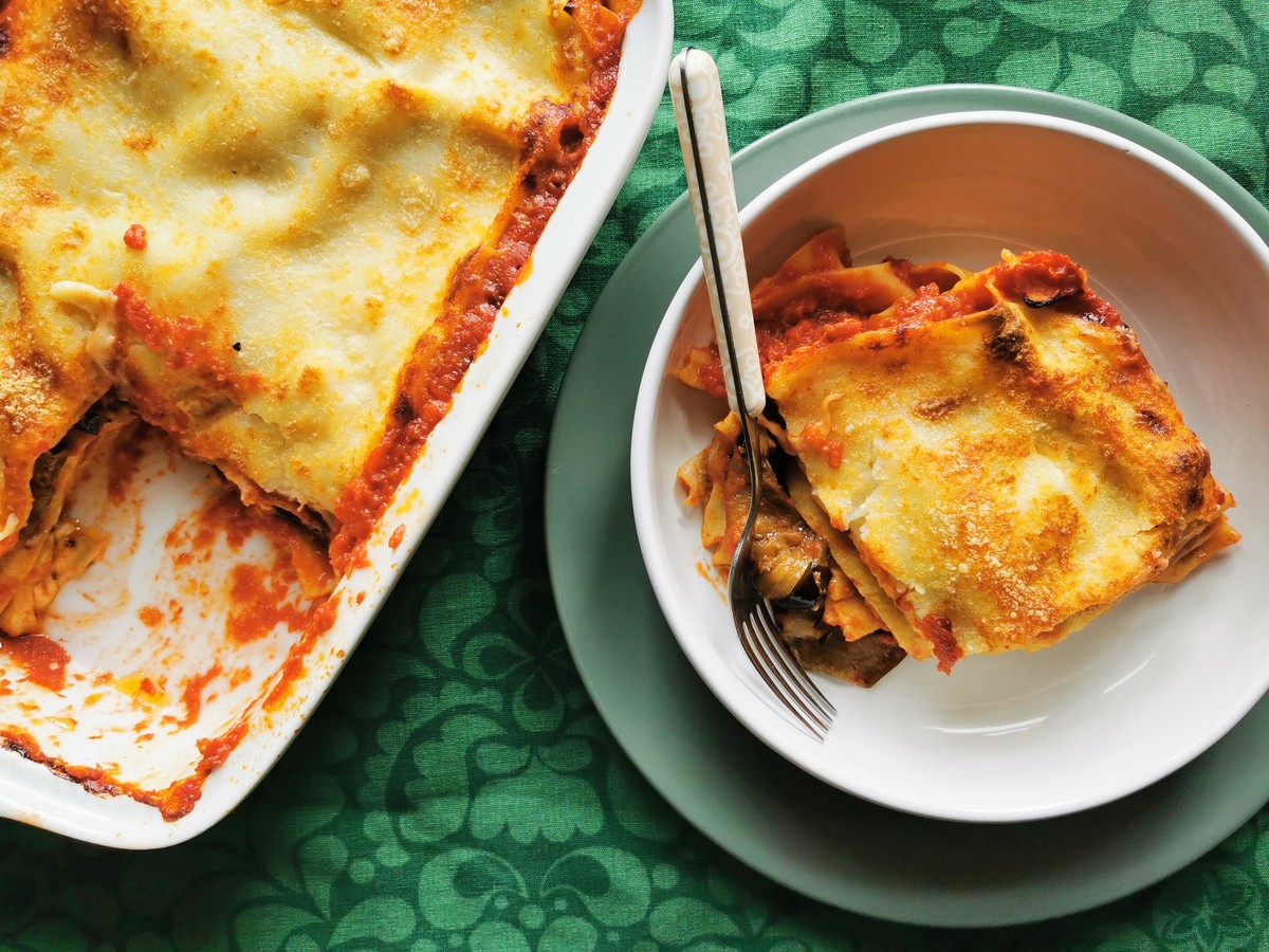 Ready baked eggplant parmigiana lasagna in oven dish with one portion in white bowl.