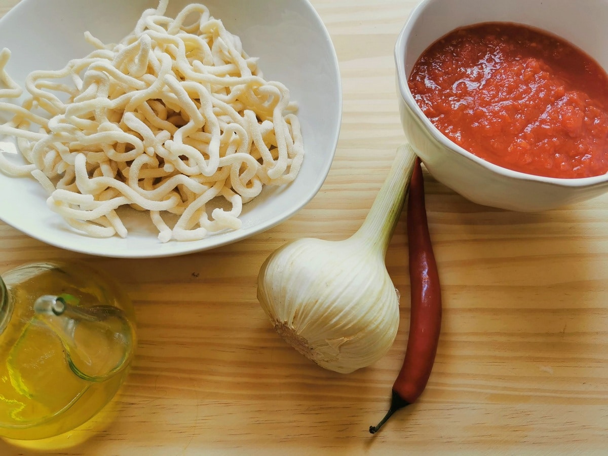 Ingredients for pici all'aglione on wood surface; pasta, tomato passata, elephant garlic, peperoncino and olive oil.