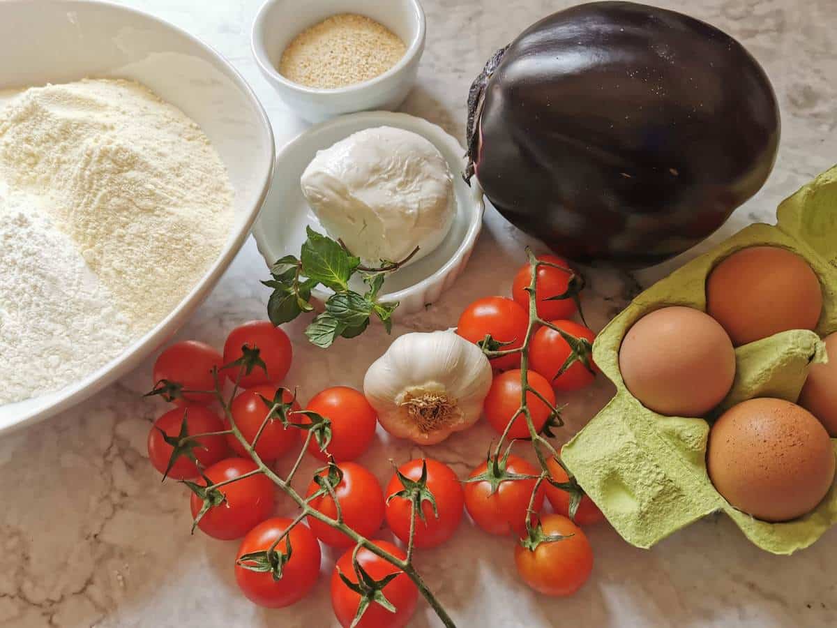 Eggs, durum wheat semolina flour, Italian 00 flour, eggplant, fior di latte mozzarella, breadcrumbs, fresh mint and cherry tomatoes on a kitchen countertop.