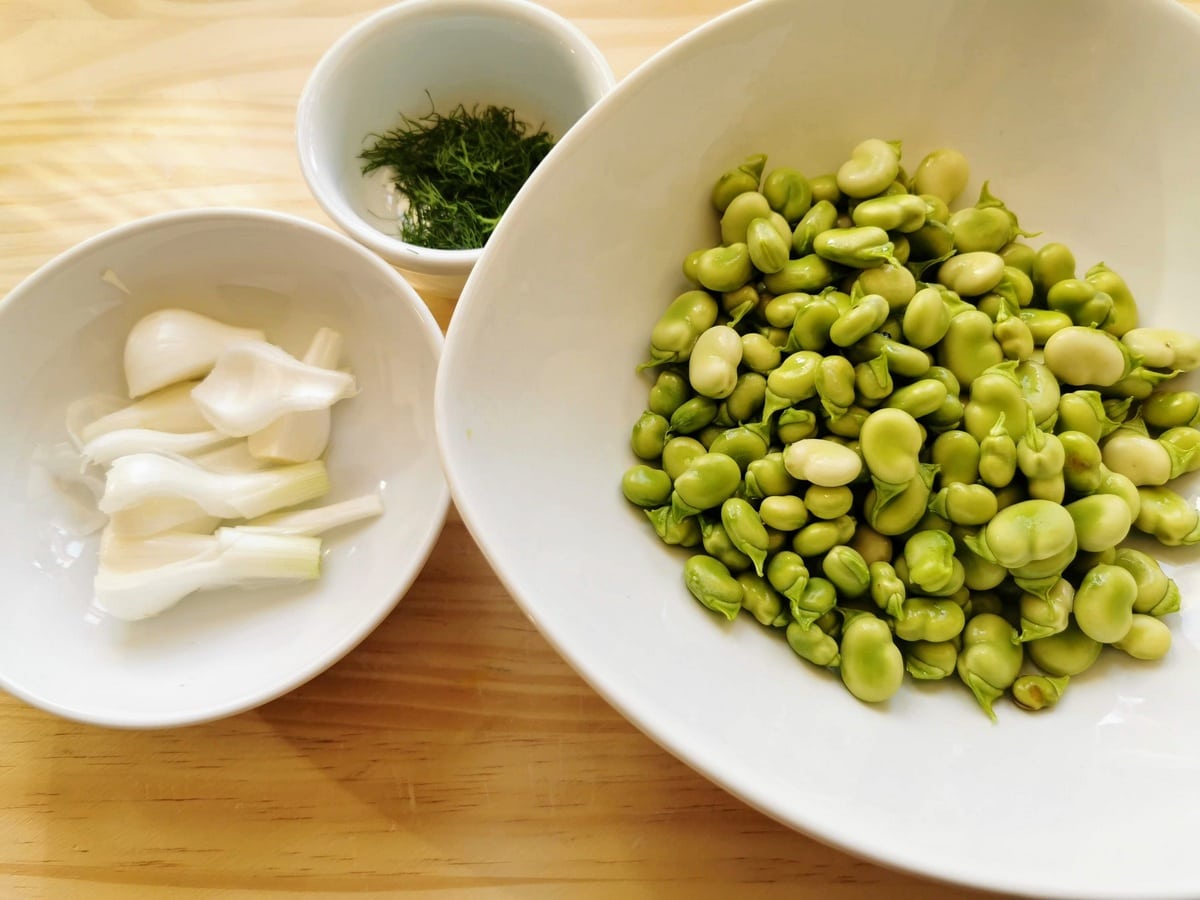 Shelled fava beans in white bowl with peeled and chopped spring onion in white bowl and chopped wild fennel in white bowl