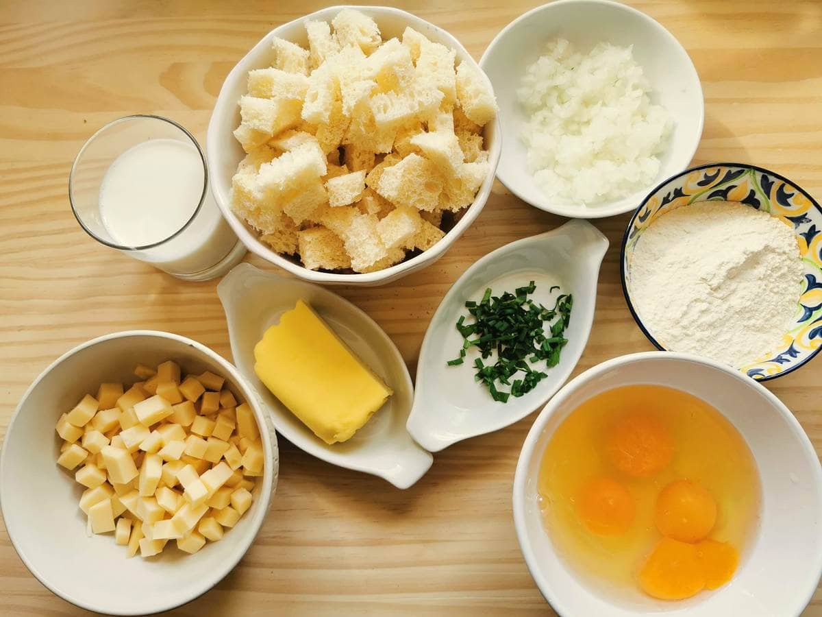 Prepared ingredients for fried canederli, stale bread cubes, chopped onion, cubed cheese, open eggs, flour, butter and chopped chives in small bowls.