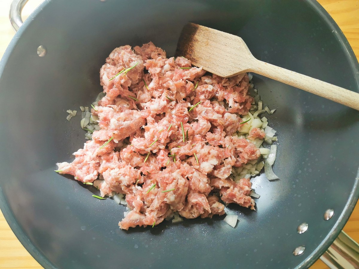 Raw sausage meat in skillet with sauéed onions and fresh rosemary leaves.