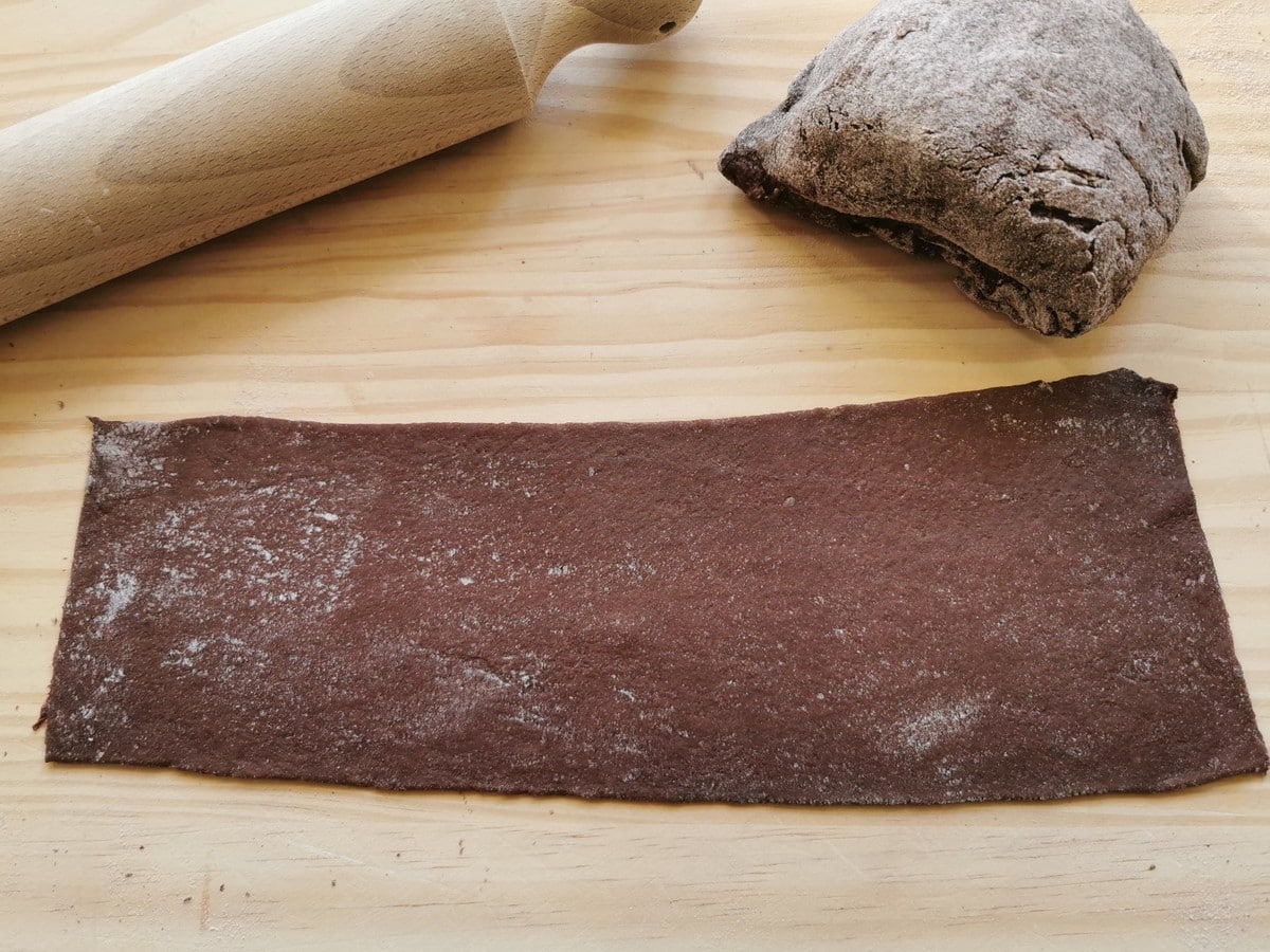 A sheet of chocolate ravioli dough on a wood work surface next to a rolling pin.