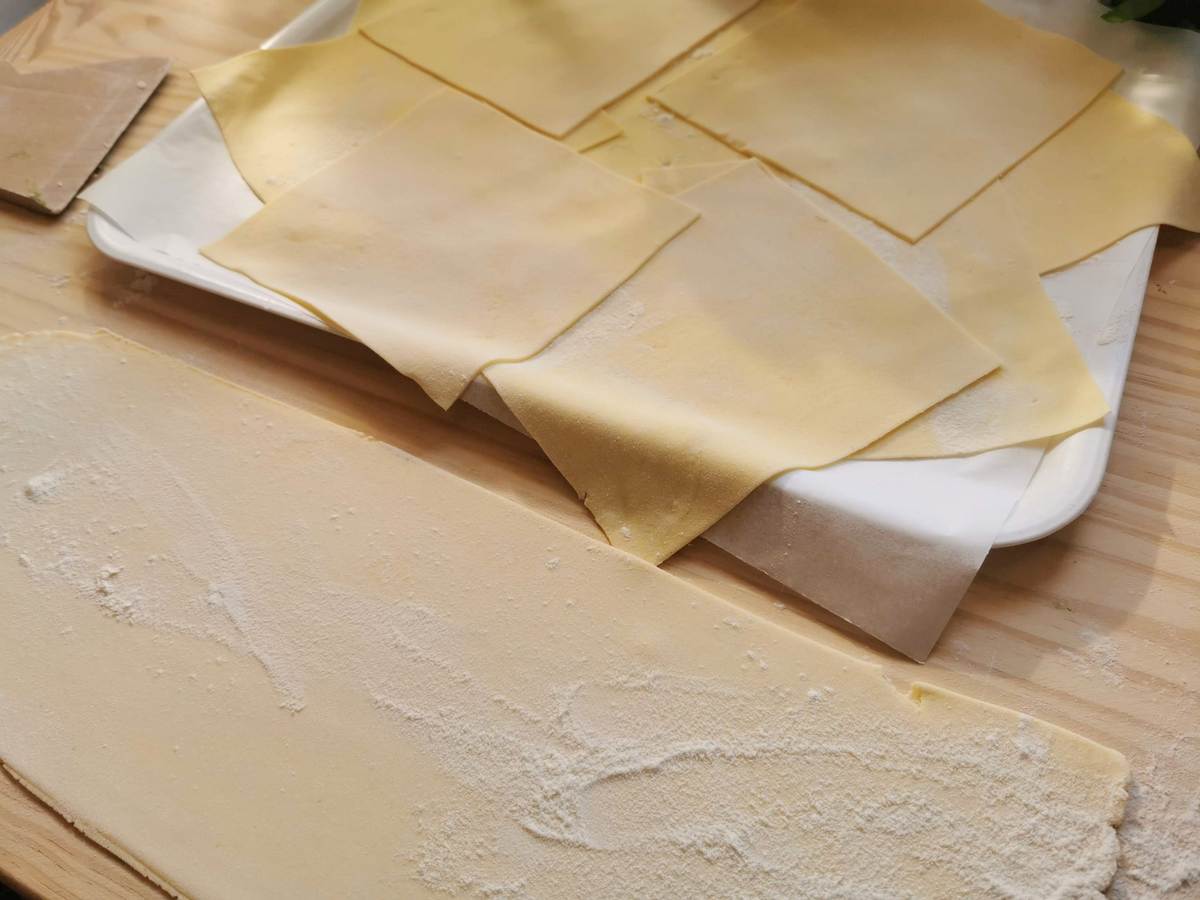 fresh pasta dough squares on white tray and pasta sheet on wood pastry board