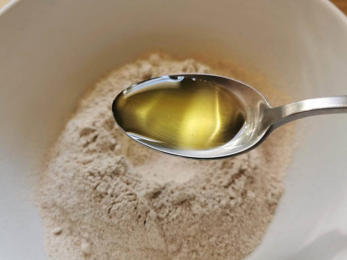 A tablespoon of olive oil held over a white bowl with flour in it.