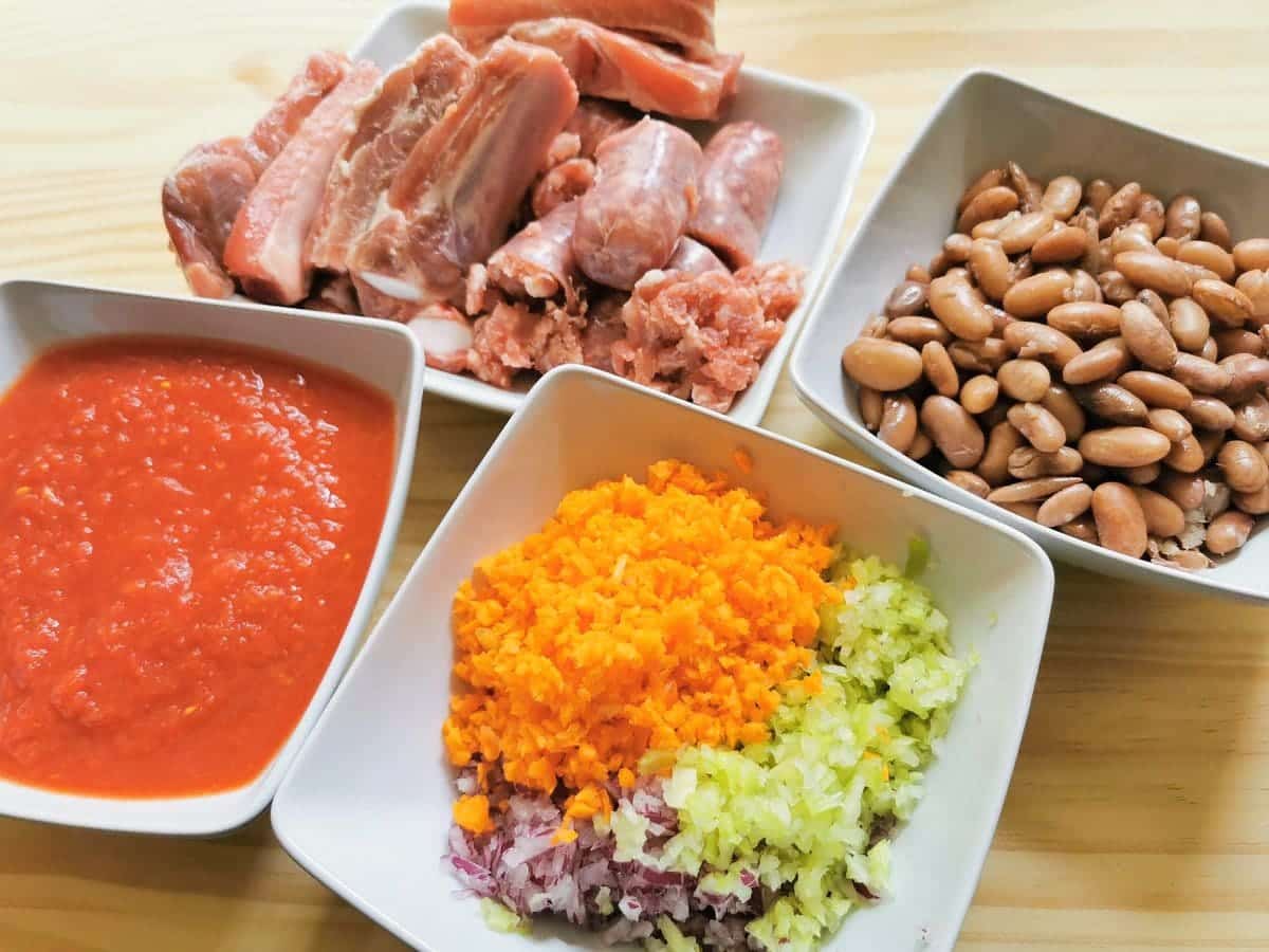 cooked borlotti beans, tomato passata, sausages and pork ribs, chopped onion, celery and carrots all in separate white bowls.