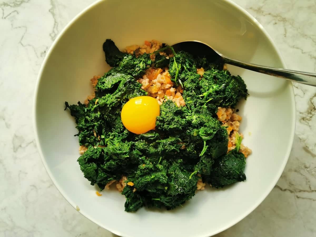 cooked meat, chopped spinach and a raw egg in a large white bowl.