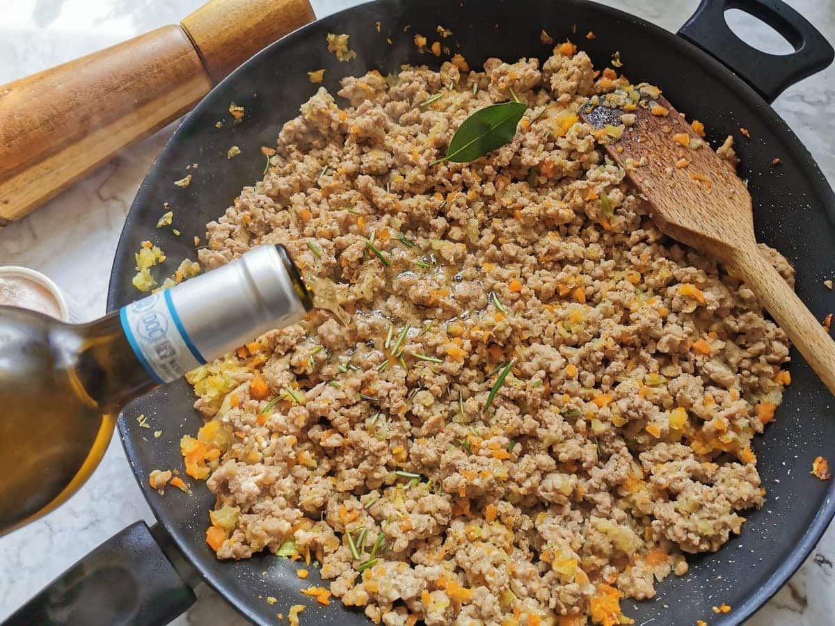 Wine being added to cooked ground meat, vegetables and herbs in large frying pan.