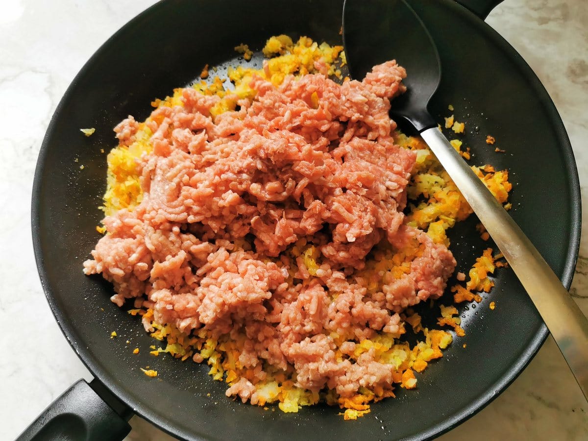 Sautéed onion, carrot and celery in frying pan with uncooked ground pork and veal on top.