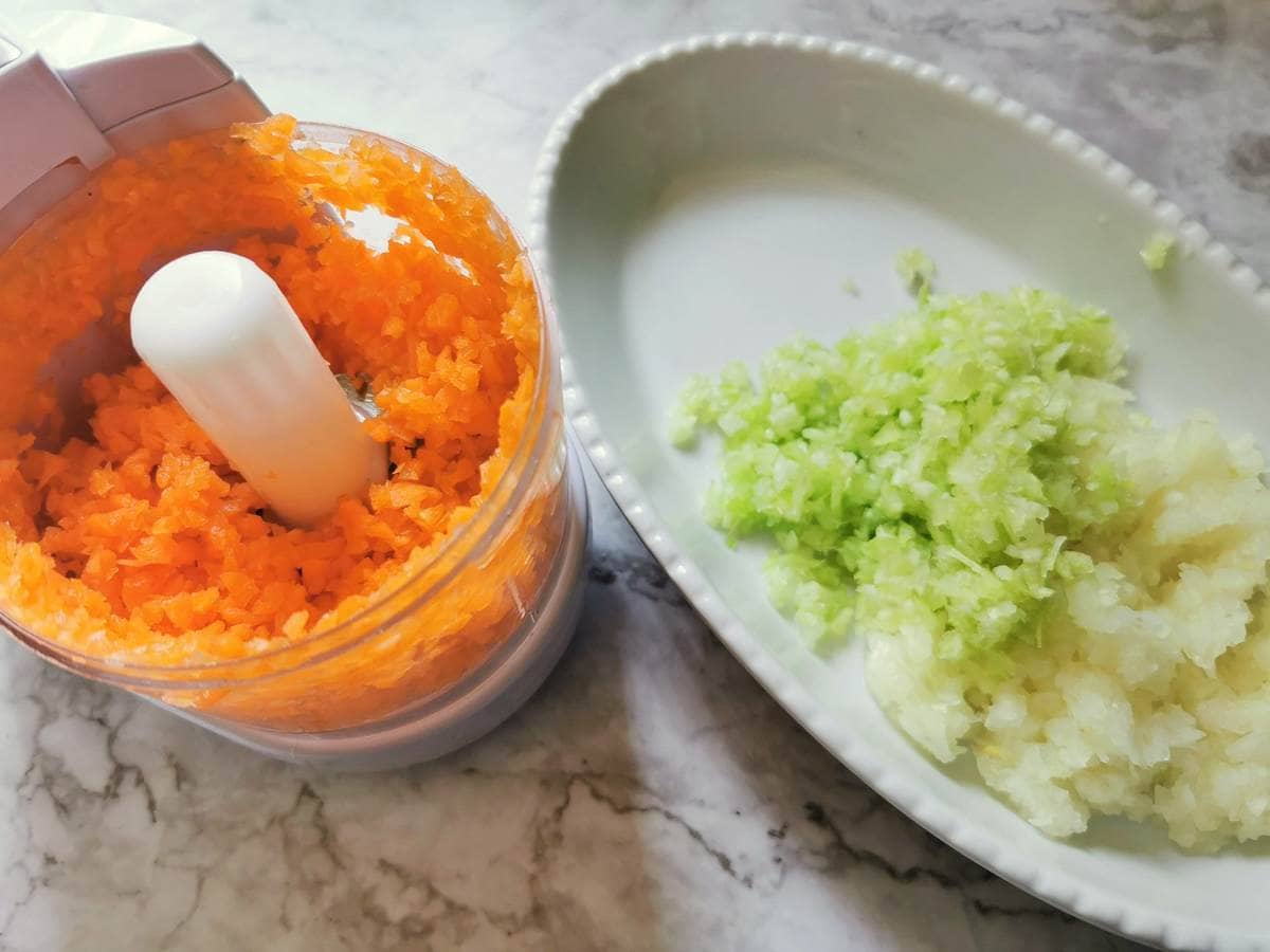 Finely chopped onion and celery in oval white bowl. Finely chopped carrot in food processor.