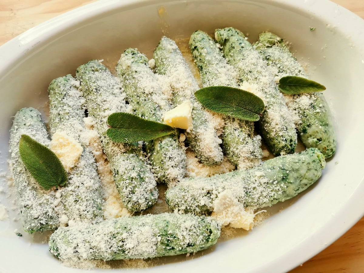 Boiled spinach gnocchi covered in grated parmigiano and some sage leaves in oven dish.