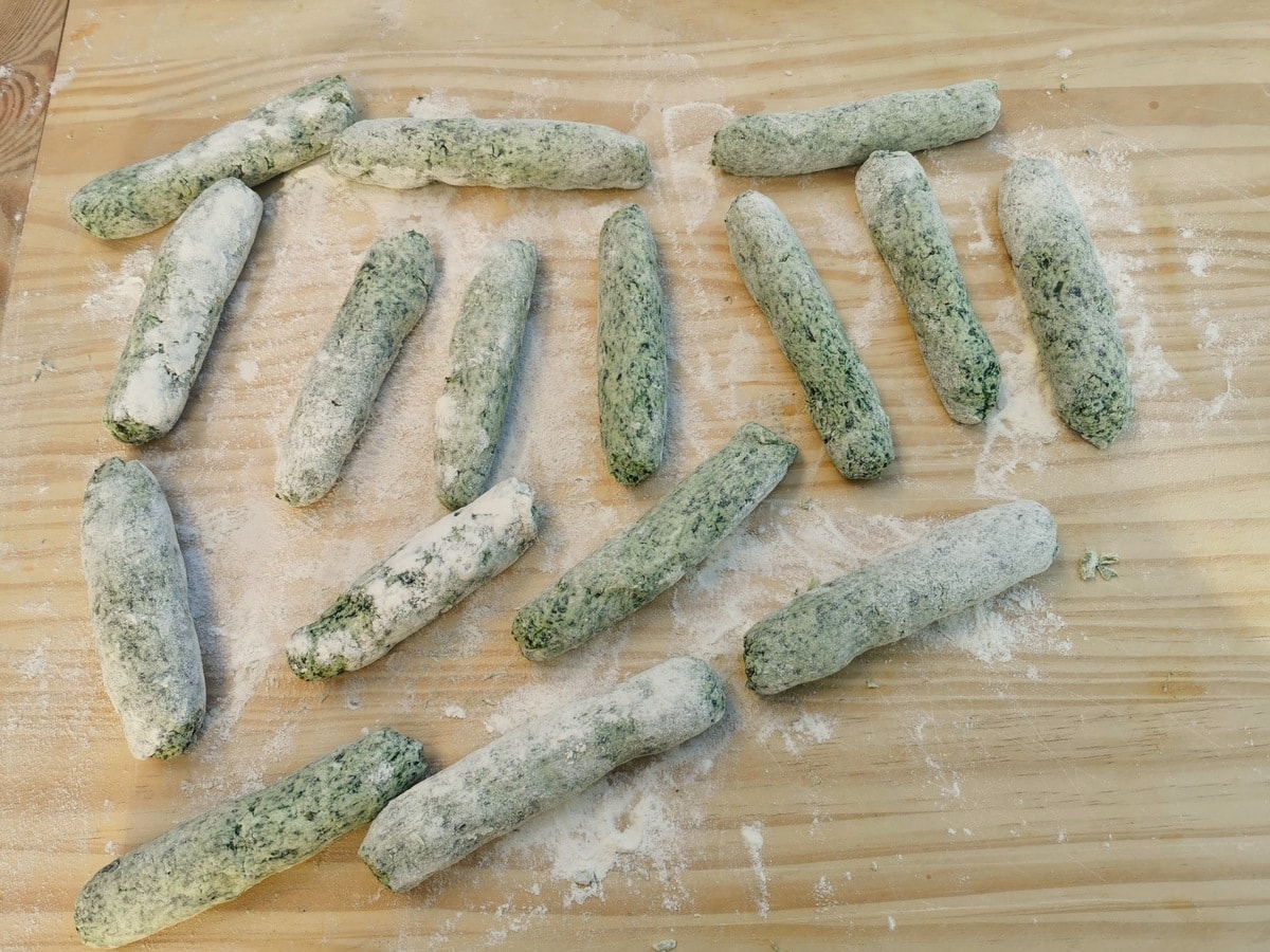 Uncooked large cigar shaped spinach ricotta gnocchi on wood pastry board.