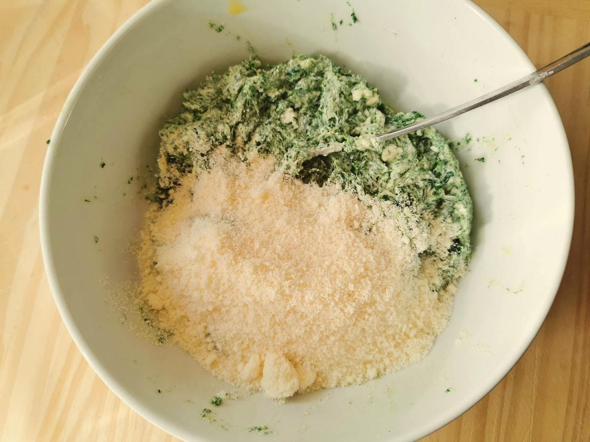 Grated Parmigiano cheese in white bowl with spinach gnocchi mixture.