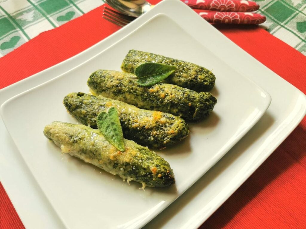 A square white plate displays four baked zucchinis coated in cheese and herbs, garnished with fresh sage leaves. The plate is set on a red placemat on top of a green and white checkered tablecloth. In the background, there are red floral-patterned napkins and a spoon.