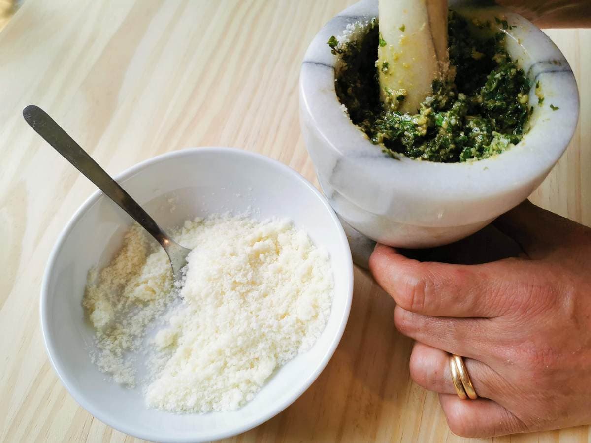 grated cheese being added to mashed basil, garlic and pine nuts using a pestle and mortar