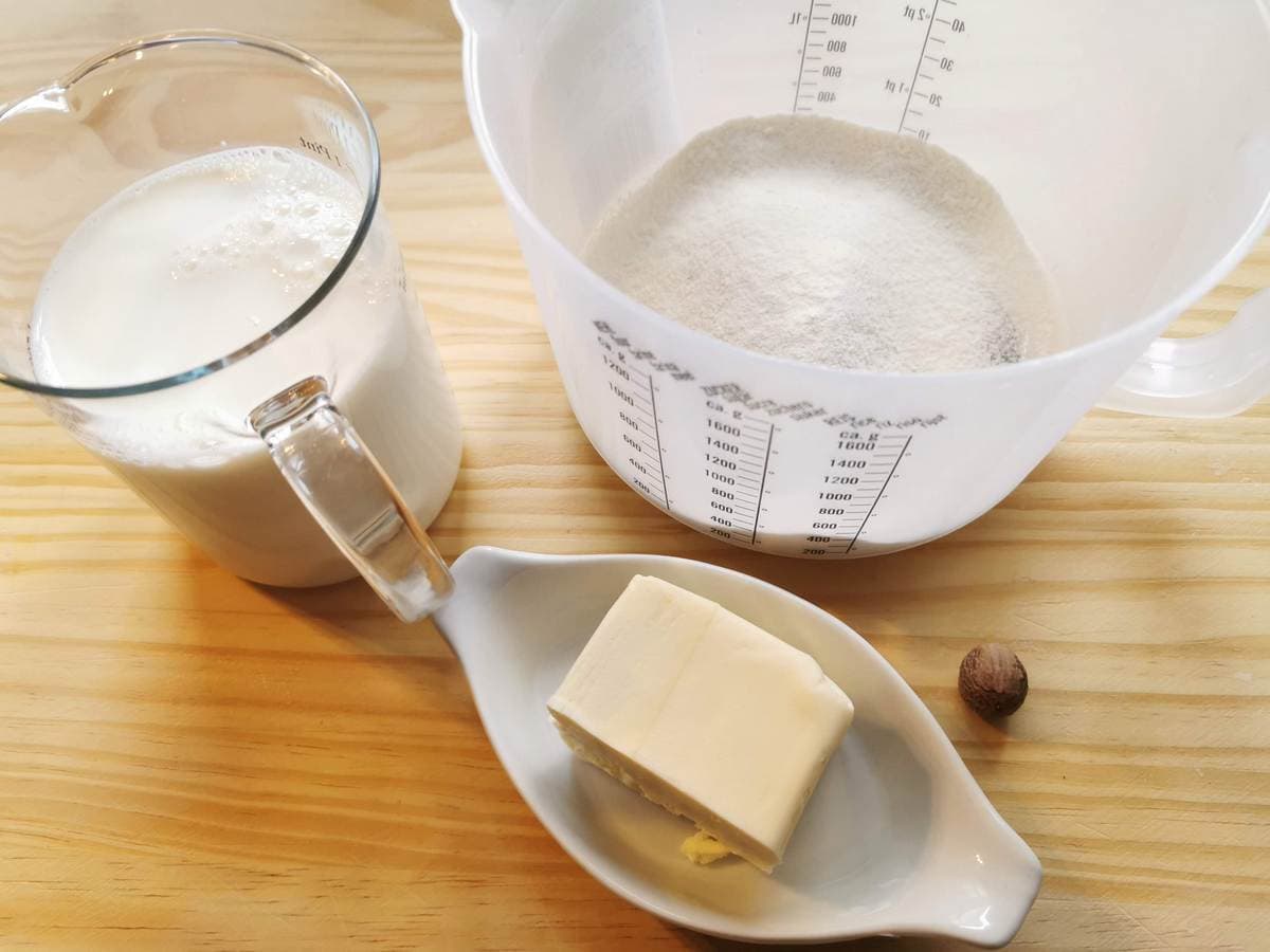 A small jug of milk, a large pat of butter, nutmeg and a plastic measuring jug with flour on wood work surface.